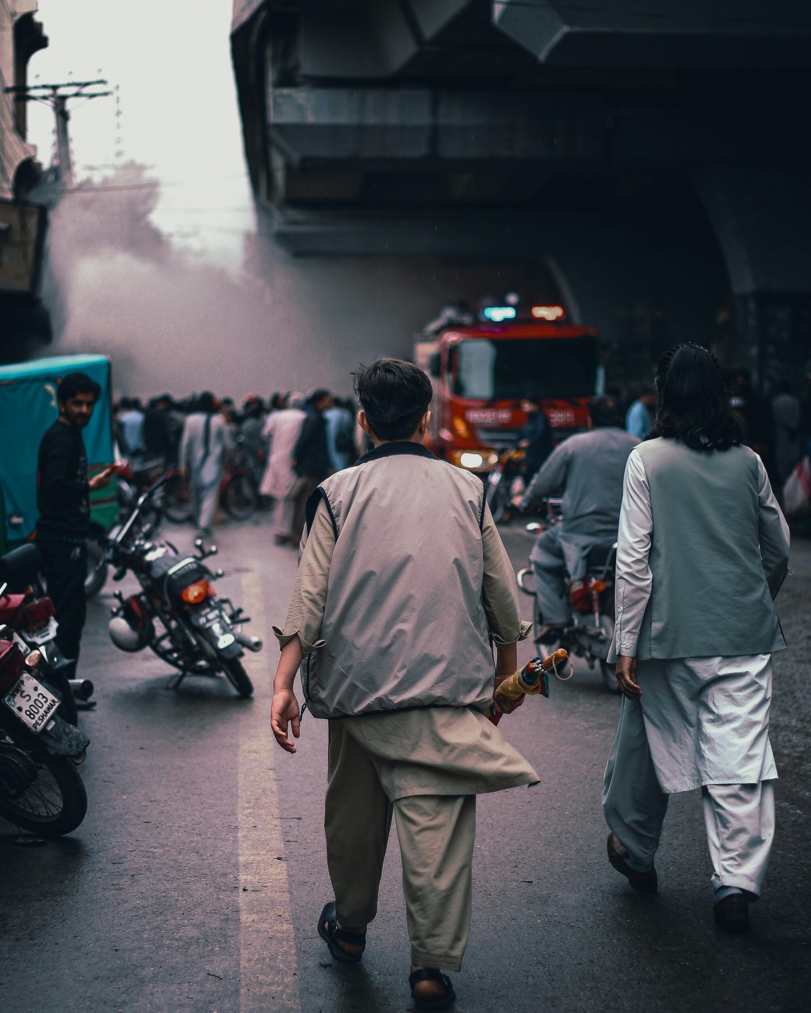 Firefighters at an emergency scene in Peshawar, Pakistan. Crowd and motorcycles surround the area under a bridge.